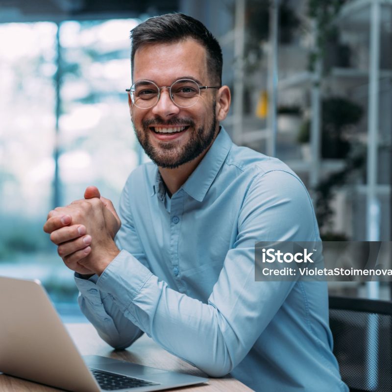 Shot of a businessman sitting on desk in modern office and working on laptop. Elegant handsome businessman sitting in his office and using laptop.